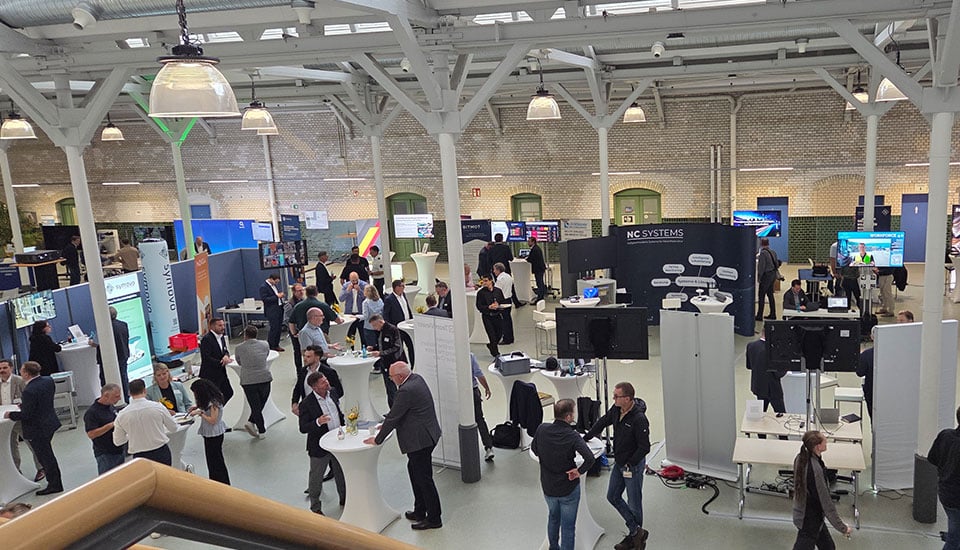 Blick in die Ausstellungshalle der 5G-Fachtagung bei Infraserv Höchst mit zahlreichen Messeständen, Besuchern und Gesprächsrunden.