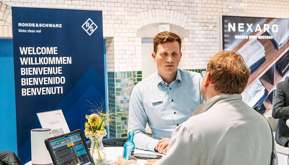 Stand von Rohde & Schwarz auf der 5G-Fachtagung: Mitarbeiter im Gespräch mit Besucher, im Hintergrund Banner von Rohde & Schwarz.