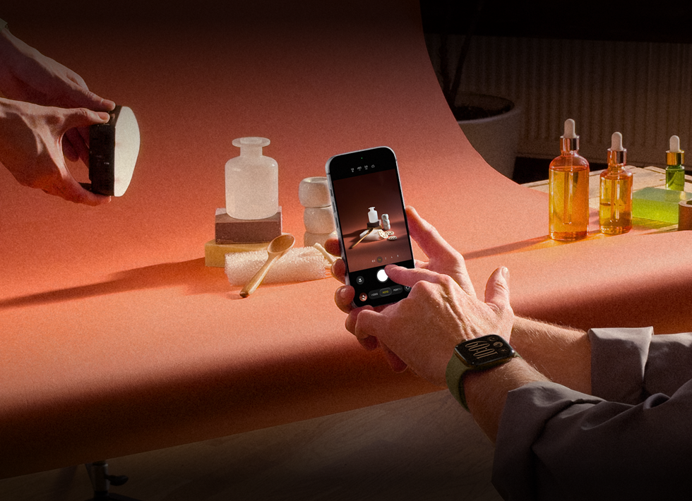 Ein Fotoshooting an einem Arbeitsplatz, eine Person fotografiert mit einem iPhone Produkte vor einem Hintergrund, eine Person hält eine Lampe