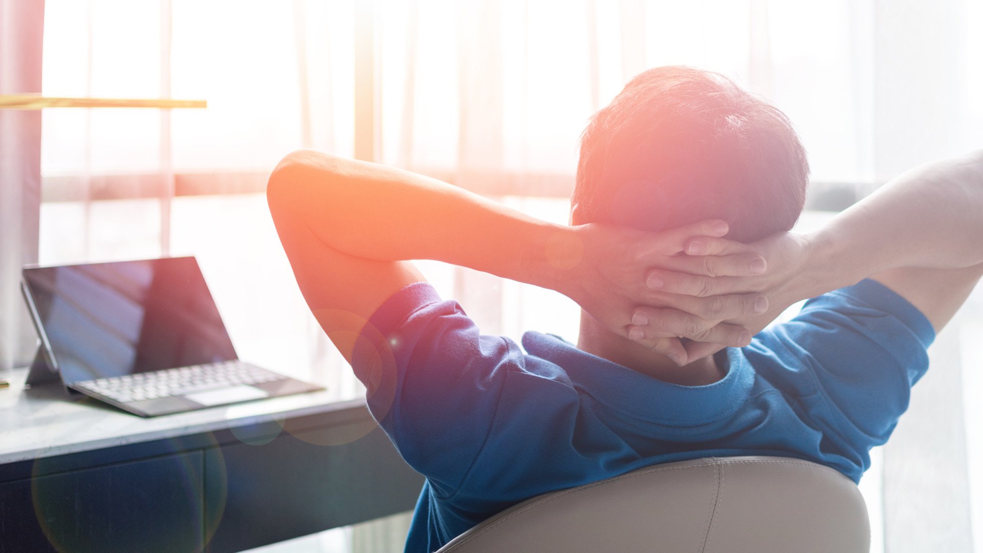 Ein Mann sitzt mit hinter dem Kopf verschrenkten Armen am Schreibtisch und schaut aus dem Fenster.