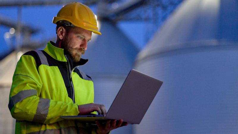 Ein Mann in Arbeitskleidung tippt auf einem Laptop vor einer Fabrikanlage