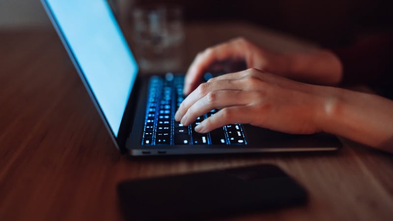 Nahaufnahme weiblicher Hände, die auf einem Laptop tippen. Abgedunkelter Raum, einzige Lichtquelle sind Tastatur und Bildschirm des Laptops.