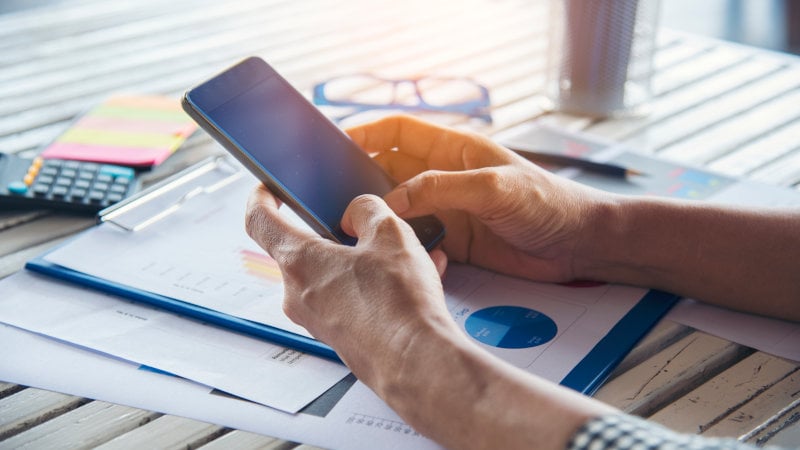 Eine Frau hält ein Smartphone in den Händen während sie über Unterlagen sitzt