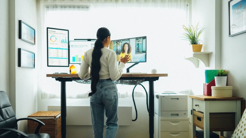 Junge Frau arbeitet im Homeoffice an einem Stehschreibtisch mit mehreren Monitoren und geht dabei auf einem Laufband.