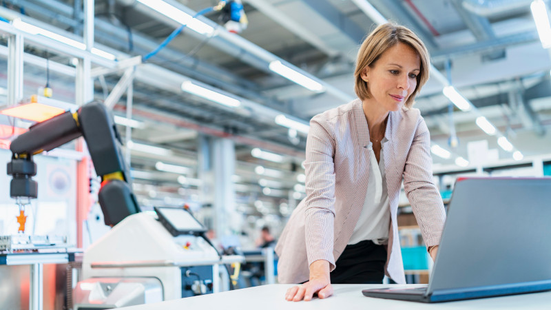 Eine Geschäftsfrau schaut in einer Fabrikhalle auf einen Laptop. Im Hintergrund befindet sich eine Produktionsstraße mit Roboter.