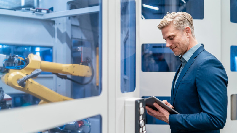 Ein Mann im Anzug steht mit einem Tablet vor einem Roboter in einer modernen Fabrik