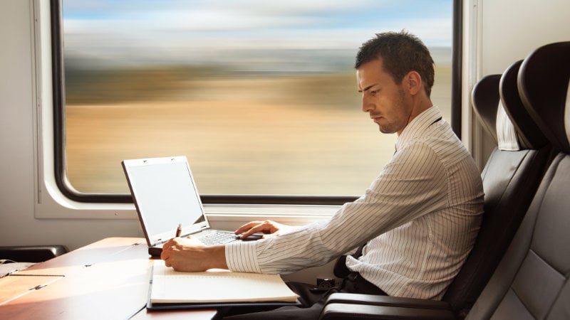 Junger Mann sitzt im Zug und arbeitet am Laptop.