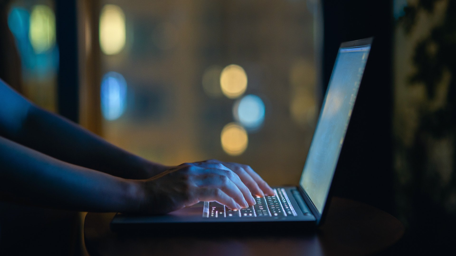 Zwei Hände tippen auf beleuchteter Tastatur von einem Laptop vor verschwommenem dunklen Hintergrund.