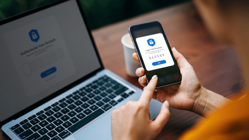 Eine Frau sitzt vor einem Laptop und nutzt Zwei-Faktor-Authentifizierung auf einem Handy