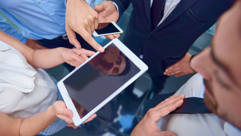 Gruppe von Leuten in Businesskleidung schaut zusammen auf Tablet in der Mitte