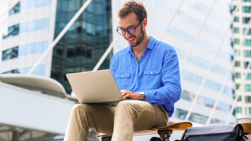 Abbildung: Ein lächelnder junger Mann mit Brille sitzt im Freien auf einem Skateboard und arbeitet auf einem Laptop; im Hintergrund sind moderne Glasgebäude zu sehen.