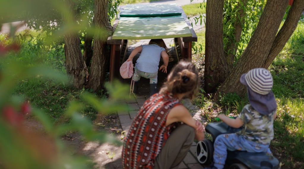 Abbildung: Junge Frau sitzt in der Hocke auf einem Weg und schaut Kindern beim Spielen zu.