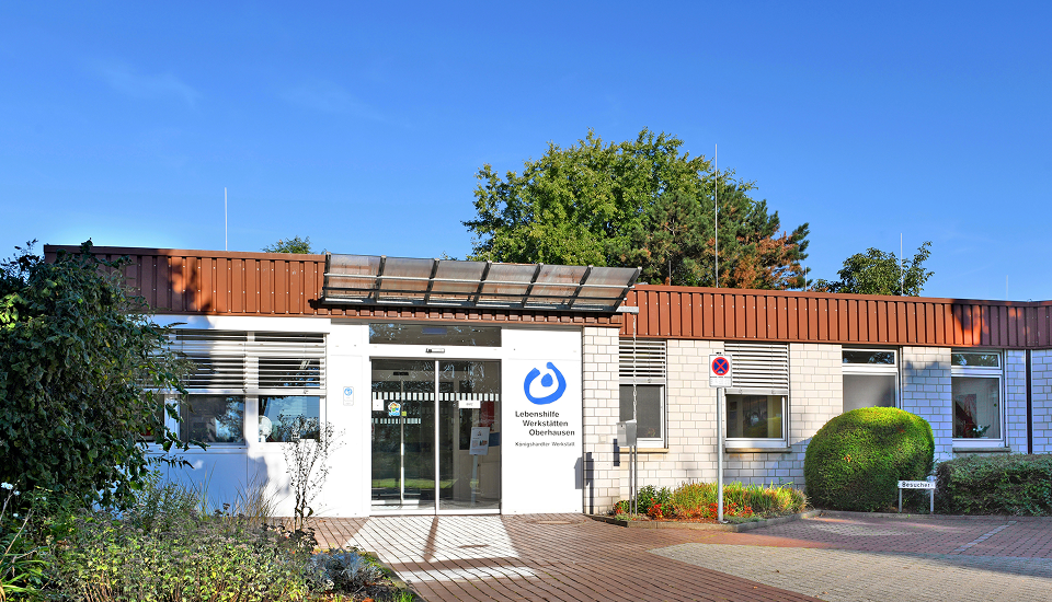 Eingangsbereich der Lebenshilfe Werkstätten Oberhausen mit modernem Flachbau, gläserner Eingangstür und gepflegter Grünanlage.