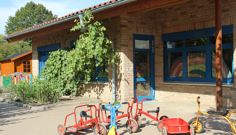 Außenansicht eines Kindergartengebäudes mit Backsteinfassade, blauen Fenstern und mehreren Kinderfahrzeugen auf dem Hof vor dem Eingang.