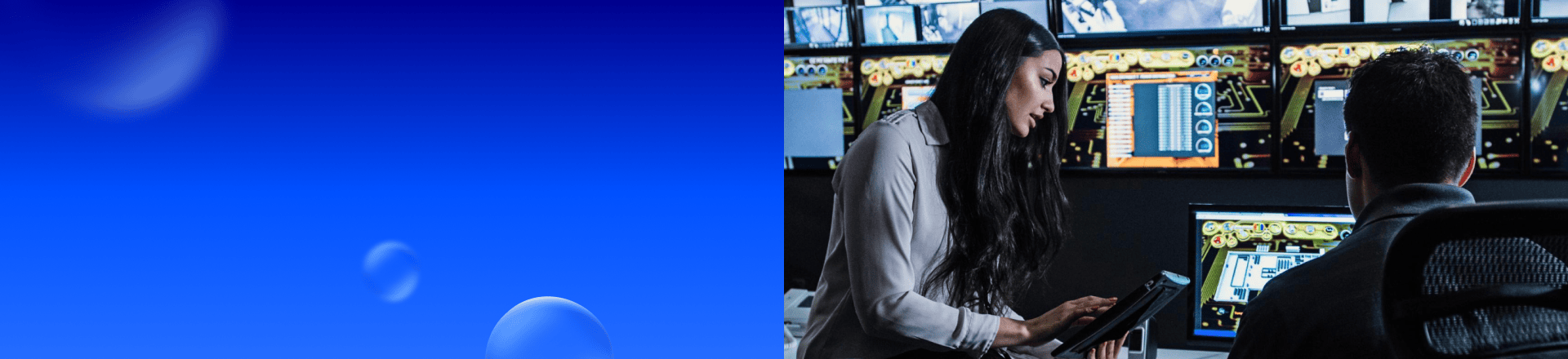 Frau mit Tablet in einer Cybersecurity-Leitstelle neben einem sitzenden Mann; im Hintergrund zahlreiche Monitore mit technischen Anzeigen.