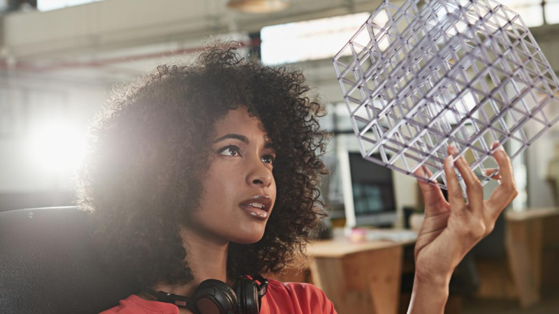 Abbildung: Eine junge Frau sitzt in einem modernen Büro und hält ein geometrisches 3D-Modell aus transparentem Kunststoff in der rechten Hand, als würde sie es prüfen.  Sie trägt ein rotes Oberteil und hat dunkles, voluminöses, lockiges Haar in mittlerer Länge. Um den Hals hängt ein schwarzes Over-Ear-Headset.  Im Hintergrund sind Schreibtische, Computer und helle Fenster zu sehen. Die Szene vermittelt einen kreativen, technischen Arbeitskontext.