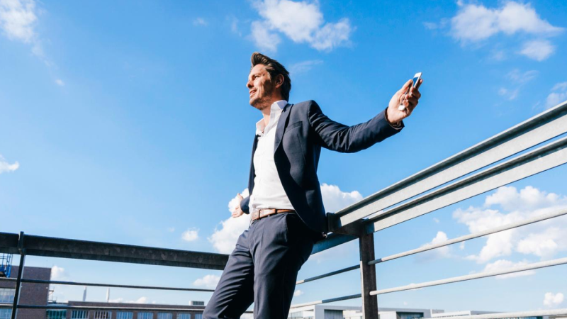 Abbildung: Ein junger Erwachsener (männlich) steht auf einer Dachterrasse oder einem erhöhten Außenbereich vor einem Geländer unter blauem Himmel mit vereinzelten Wolken.  Die Person trägt einen dunklen Anzug mit weißem Hemd, das oben leicht geöffnet ist, und einen hellen Gürtel. In der rechten Hand hält die Person ein Smartphone, der linke Arm ist locker am Geländer abgestützt.  Im Hintergrund sind Gebäudeteile und die Skyline zu sehen. Die Szene vermittelt einen modernen, urbanen und professionellen Kontext.
