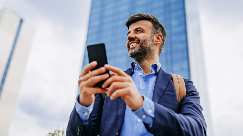 Abbildung: Ein junger Erwachsener (männlich) steht im Freien vor einem hohen, modernen Glasgebäude und hält ein Smartphone in beiden Händen.  Die Person trägt ein dunkelblaues Sakko über einem hellblauen Hemd und hat einen braunen Lederrucksack über der Schulter. Die Person schaut vom Smartphone weg und hat einen fröhlichen Gesichtsausdruck.  Im Hintergrund sind weitere Hochhausfassaden und ein heller Himmel zu sehen. Die Szene vermittelt einen urbanen, professionellen Kontext.
