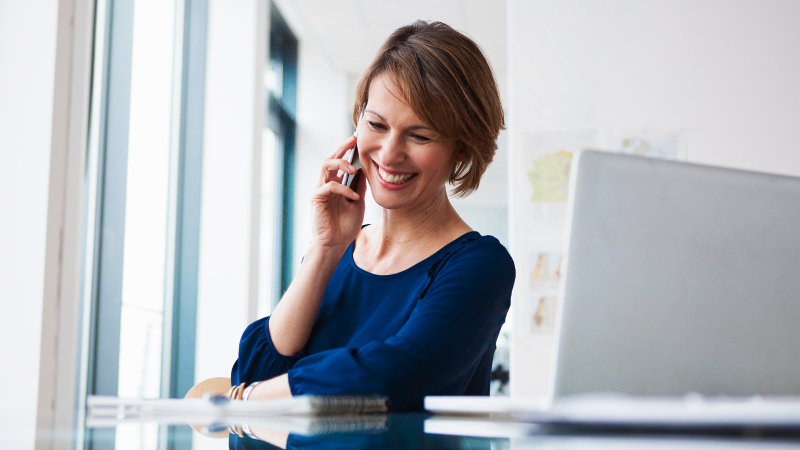 Abbildung: Eine junge Frau sitzt in einem modernen Büro an einem Tisch und telefoniert mit einem Smartphone.  Sie trägt ein dunkelblaues, langärmliges Oberteil und hat einen kurzen, gepflegten Haarschnitt. Vor ihr steht ein geöffneter Laptop, daneben liegen einige Unterlagen.  Im Hintergrund sind große Fenster mit Tageslicht und helle Wände zu sehen. Die Szene vermittelt einen professionellen, geschäftlichen Kontext.