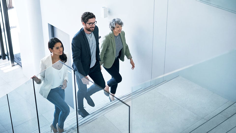 Drei Personen gehen gemeinsam eine Treppe in einem hellen Bürogebäude hinauf