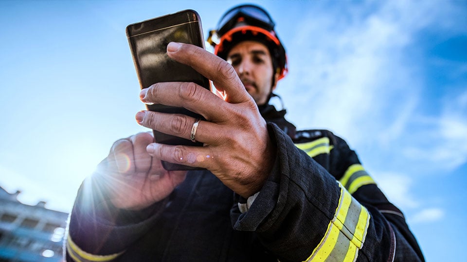 Feuerwehrmann in Einsatzkleidung mit Helm bedient ein Smartphone im Freien
